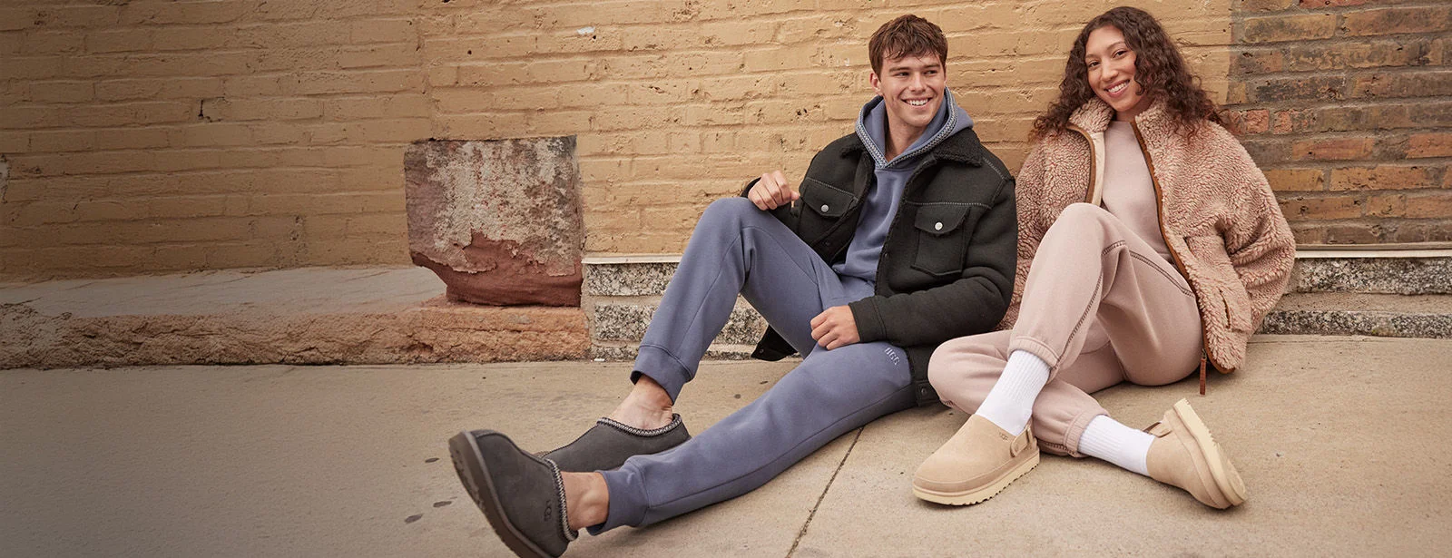 Man and woman sitting on the sidewalk wearing Uggs. 