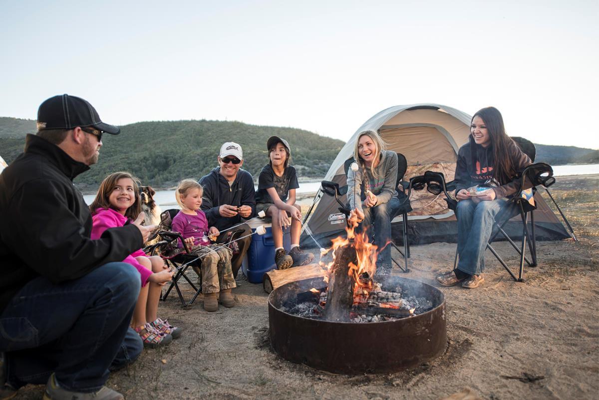 two families sitting around a campfire making smores with a lake in the background.